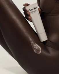 Close-up of a person holding a white VOESH product tube next to a shimmering exfoliating scrub on dark skin.