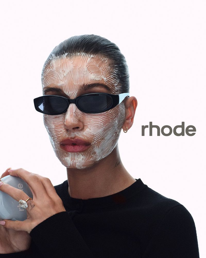 Hailey Bieber wearing sunglasses and skincare cream on her face holding a jar for rhode Skin 2026