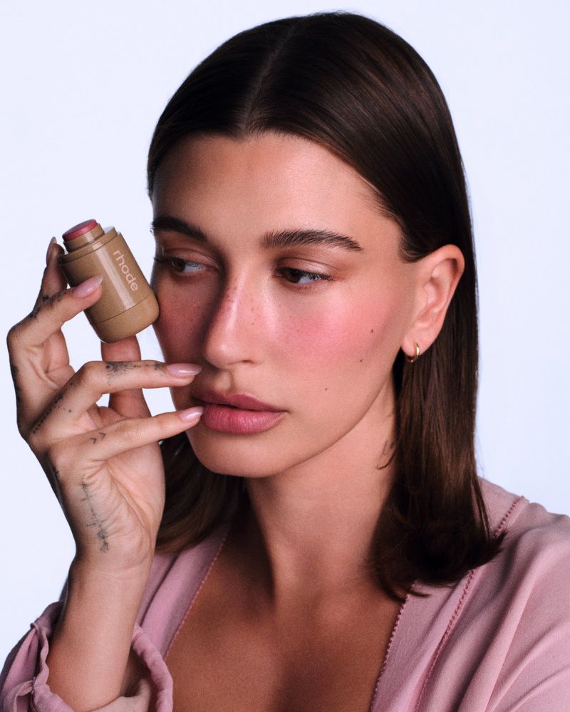Hailey Bieber holding rhode skin cosmetic product next to her cheek against a plain white background