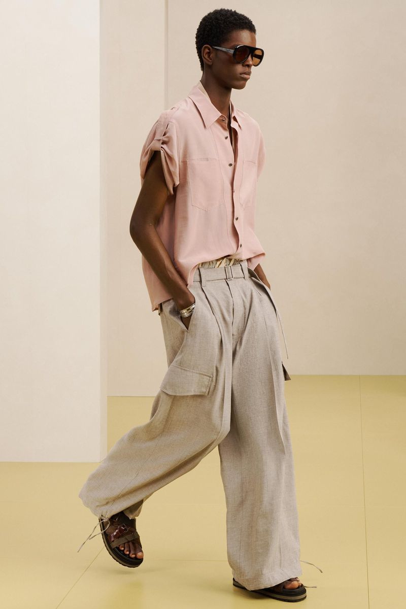 Model wearing dark sunglasses, pink short-sleeve shirt, and wide-leg trousers against a minimal studio background.