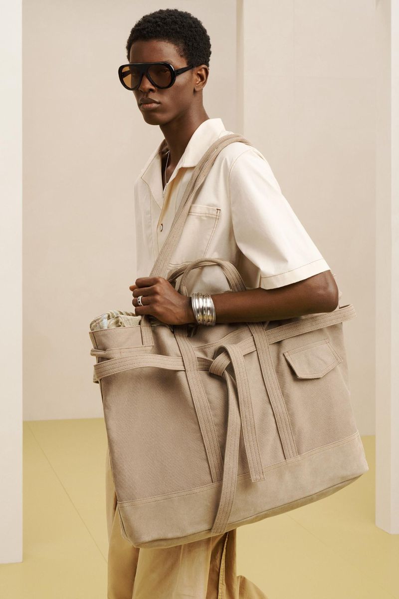 A Black male model poses in a studio with a large beige tote bag and sunglasses, wearing a light beige shirt and pants in a studio.