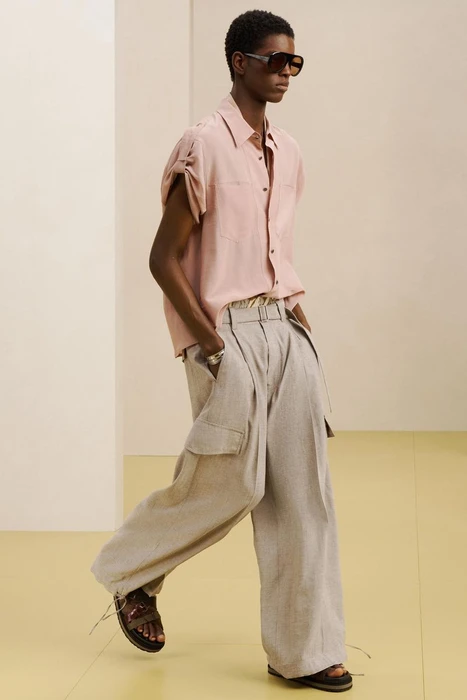 Model wearing dark sunglasses, pink short-sleeve shirt, and wide-leg trousers against a minimal studio background.