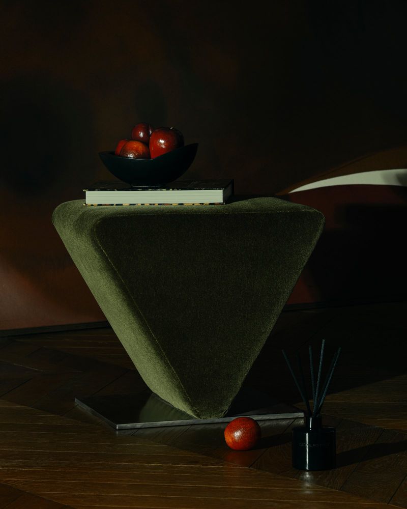Dark still life with a green velvet stool, a bowl of red fruits, a book, and a reed diffuser on a dark wood floor.