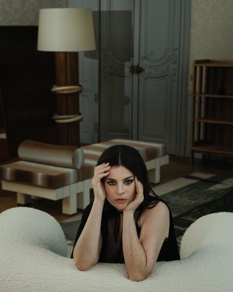 Woman with dark hair and smoky eye makeup leaning forward behind a white textured sofa in a room with a lamp and furniture