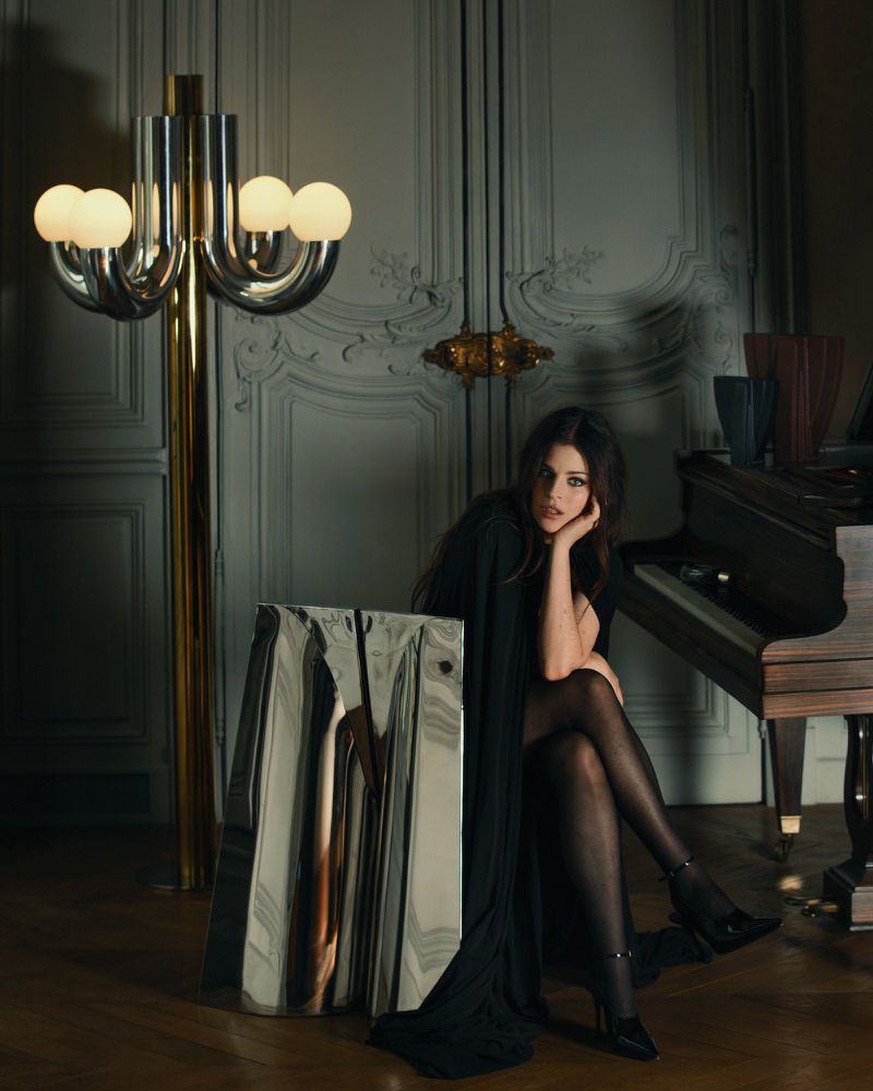 Woman in a black dress seated on a piano bench next to a sculptural floor lamp and a grand piano
