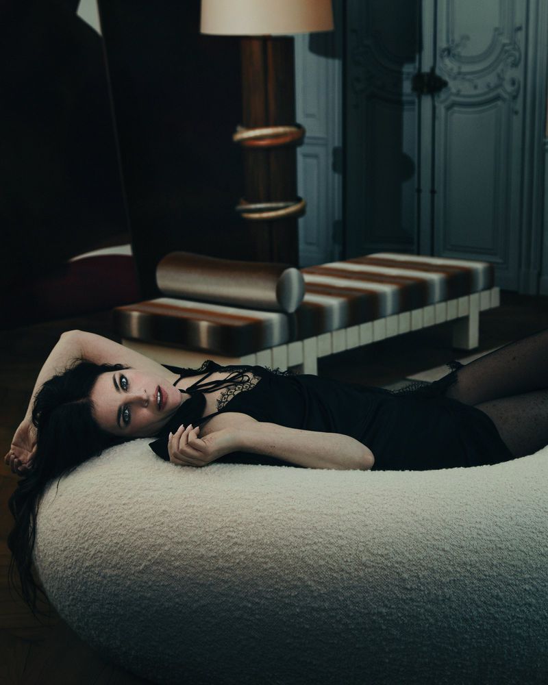 Woman with long dark hair lying on a textured white circular piece of furniture wearing a black dress