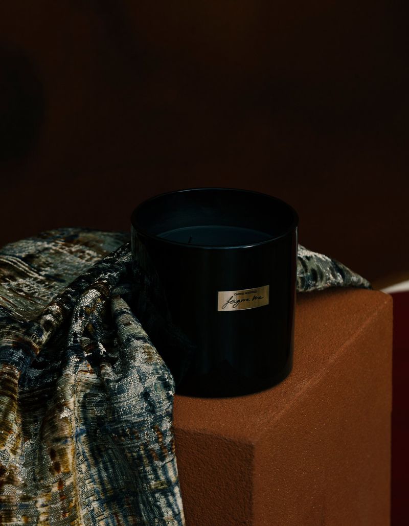 Black cylindrical candle resting on a brown pedestal next to textured patterned fabric in a dark studio setting