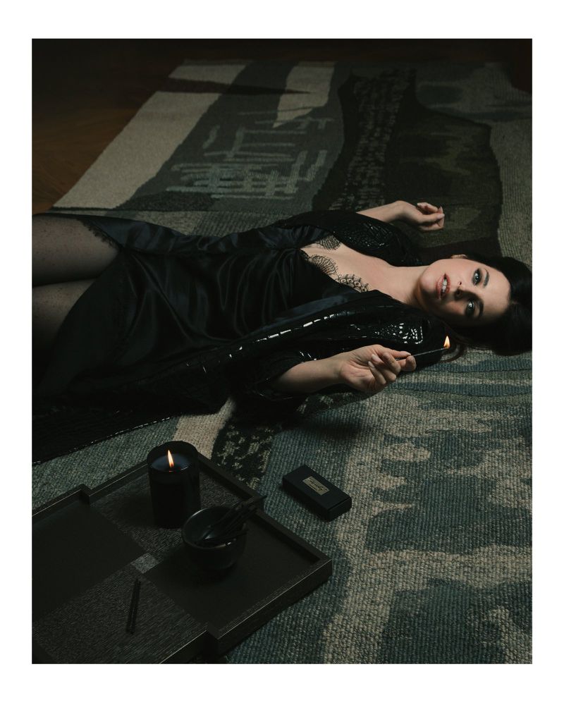 Woman in black loungewear lies on an abstract patterned rug, holding a lit match near a tray with candles in a dimly lit room.