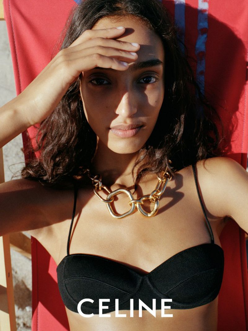 Model wearing a black bikini top and chunky gold necklace relaxing on a red sun lounger outdoors for Celine