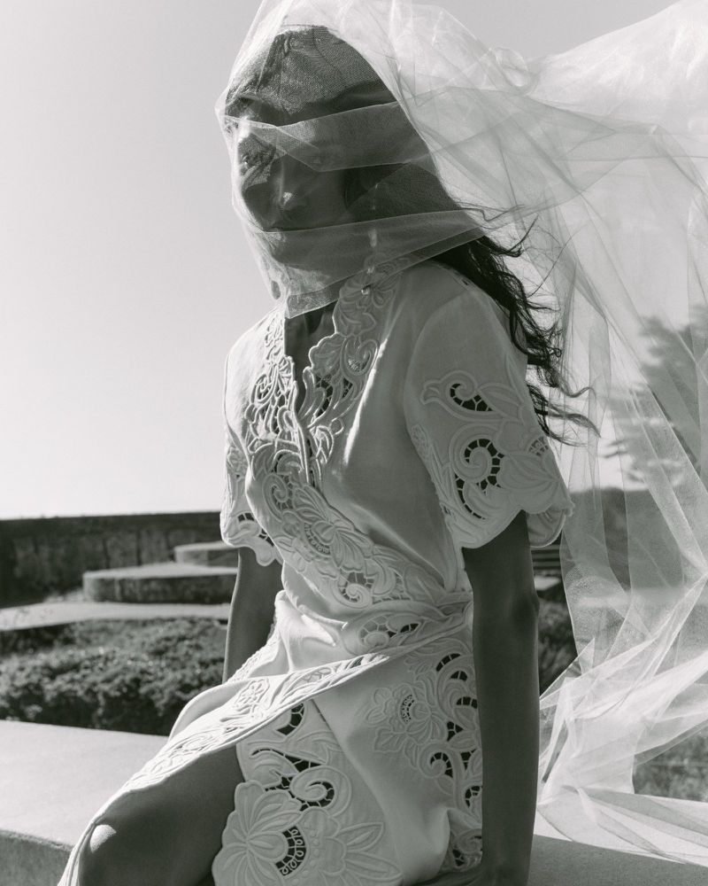 Model wearing a white lace dress with a billowing veil covering her face against an outdoor stone background