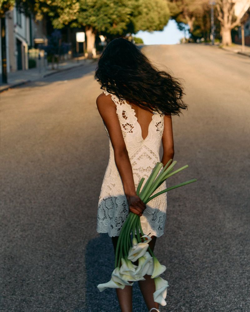 Model in a white lace dress holding a bunch of calla lilies, walking up a sunlit street for Cult Gaia.