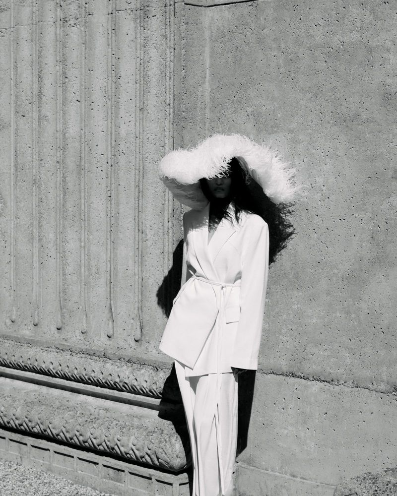 Monochrome fashion campaign image featuring a model wearing a feathered wide-brim hat and white suit against a textured stone wall