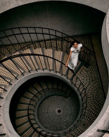 High-angle view of a model in a white dress descending a dramatic circular concrete spiral staircase with dark railings.