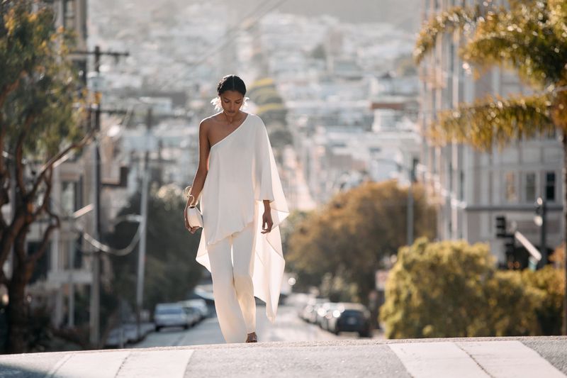 Model in a white one-shoulder flowing top and matching pants walking down a sloping city street flanked by buildings and trees.