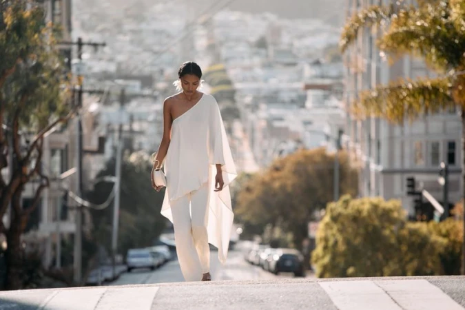 Model in a white one-shoulder flowing top and matching pants walking down a sloping city street flanked by buildings and trees.