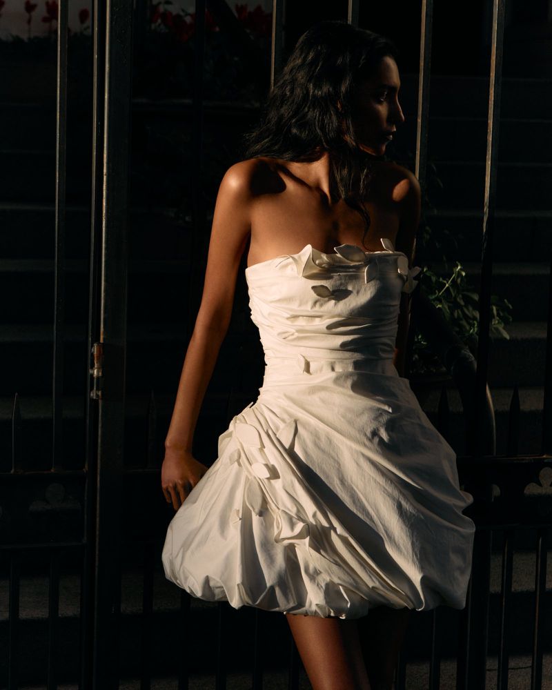 Model Shaanti Chaitram wearing a strapless white dress with petal details standing before metal gates by Arseny Jabiev for Cult Gaia.
