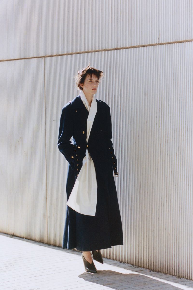 Woman in navy coat and dress with white scarf posing against a textured wall. Outdoor fashion shot with long shadows.