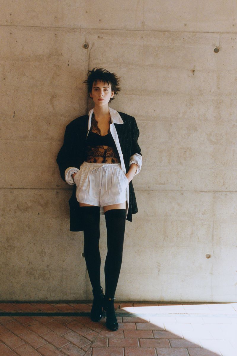 Model leaning against a textured concrete wall wearing a dark jacket, lace top, white shorts, and black thigh-high socks.