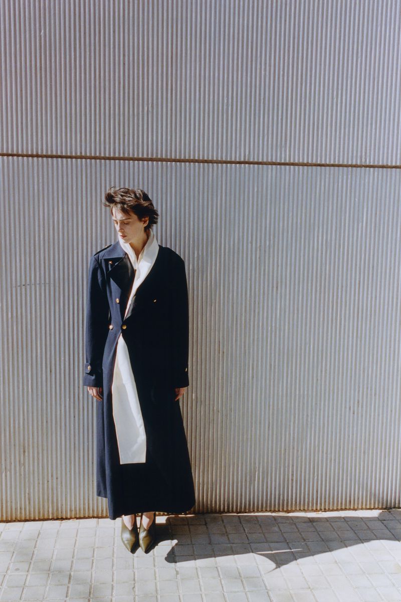 Model in a long dark coat and white scarf standing against a corrugated metal wall for Hube Magazine Raw distance