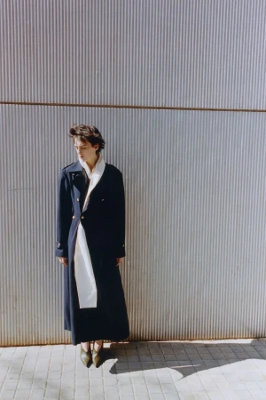 Model in a long dark coat and white scarf standing against a corrugated metal wall for Hube Magazine Raw distance