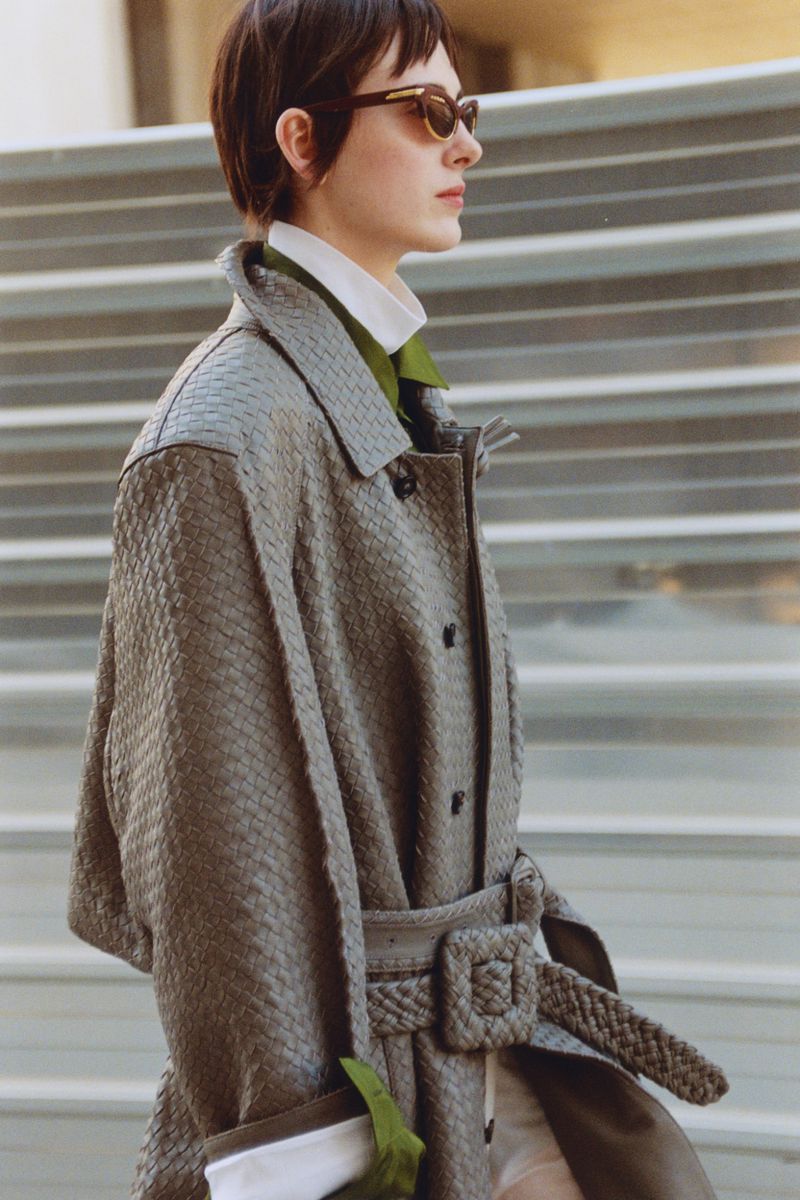 Model with dark short hair wearing sunglasses and a grey textured woven coat in profile outdoors.