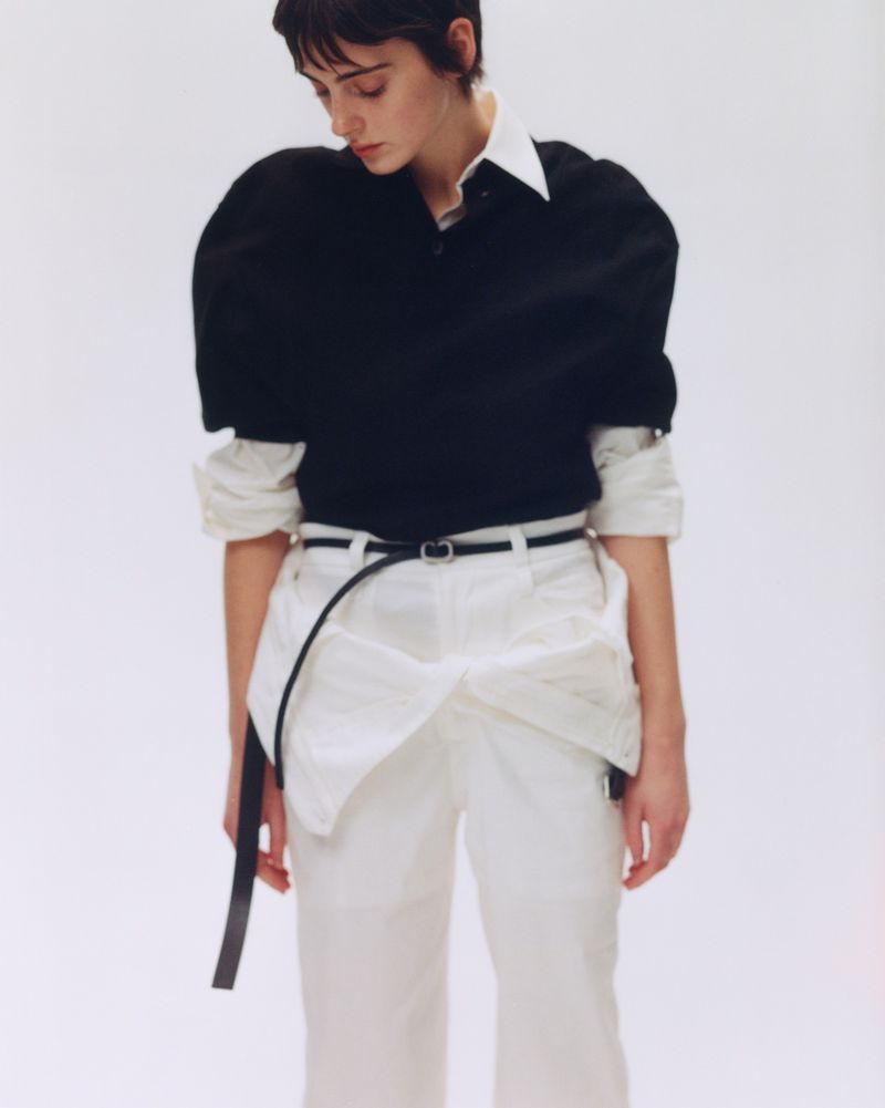 Model with short dark hair wearing a black top and white trousers against a plain white studio background