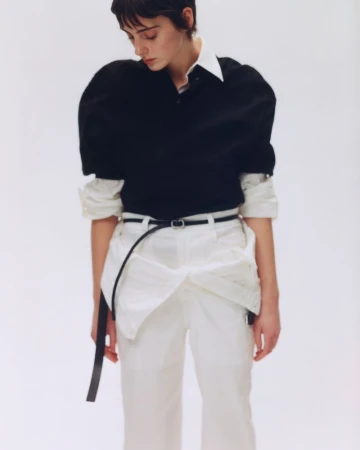 Model with short dark hair wearing a black top and white trousers against a plain white studio background