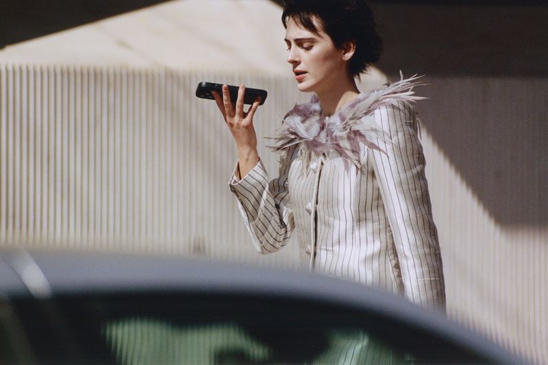 Woman in a striped jacket with feathered collar holding a phone to her ear outdoors near a car
