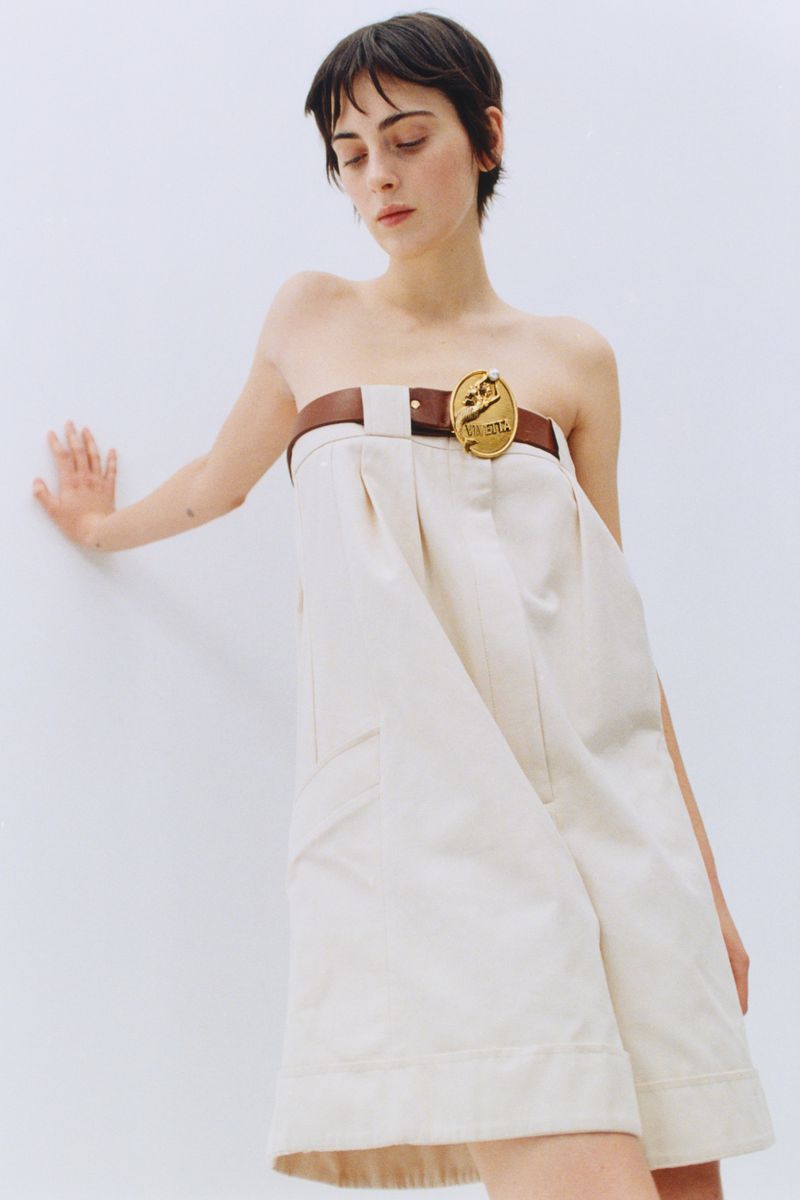 Model with short dark hair wearing an off-the-shoulder cream dress with a brown belt against a white seamless background