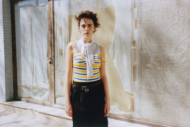 Model with short dark hair wearing a striped top and black skirt against a weathered storefront