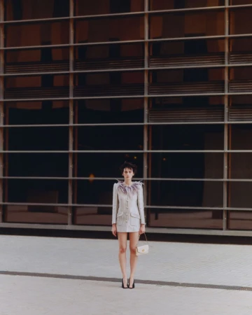 Model standing in feathered collar coat holding a small handbag in front of a modern glass building exterior