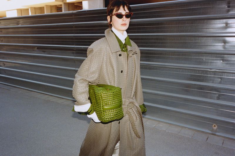 Model wearing a textured grey belted coat and green woven handbag standing outdoors in front of a corrugated metal wall.