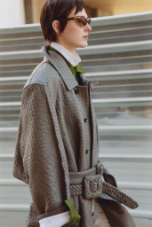 Model with dark short hair wearing sunglasses and a grey textured woven coat in profile outdoors.