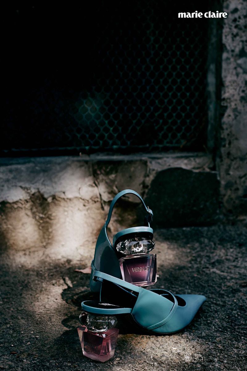 Still life product shot featuring a pair of turquoise high heel shoes and two perfume bottles on a textured concrete surface, indoors.