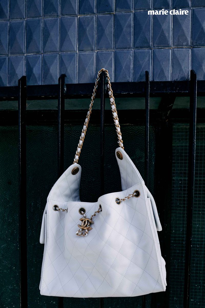 White Chanel bucket bag with gold chain strap hanging on a black iron fence with diamond patterned blue tile background, lit by natural light.