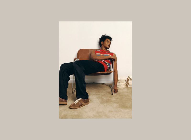 Model Nile Winchester seated in a brown leather chair wearing a red knit top and dark pants for The Webster