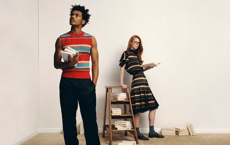 Two models standing in a studio with books, a man in a red striped vest and a woman in a striped dress beside a wooden ladder
