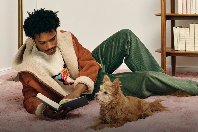 Young man wearing a brown sherpa jacket reading a book while lying on a rug, with a small dog next to him in an indoor setting.