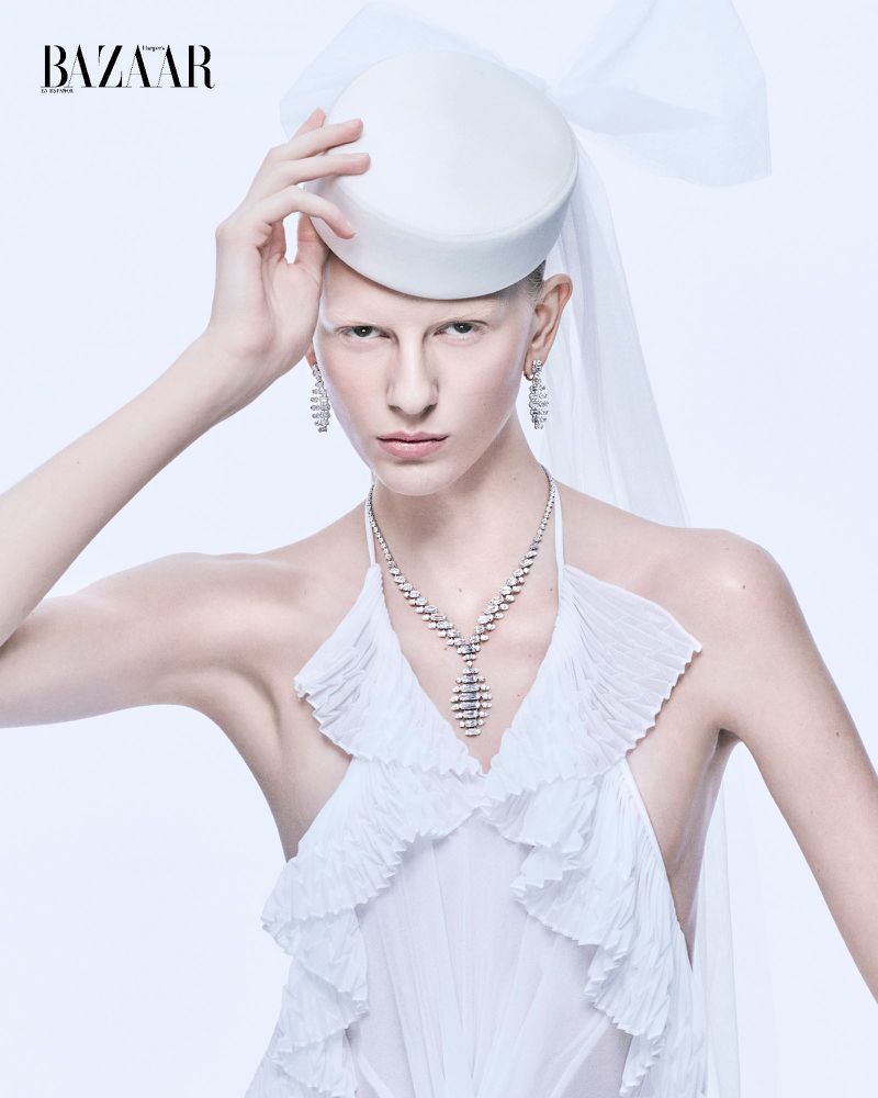 Model in white dress wearing pillbox hat and diamond necklace against white backdrop