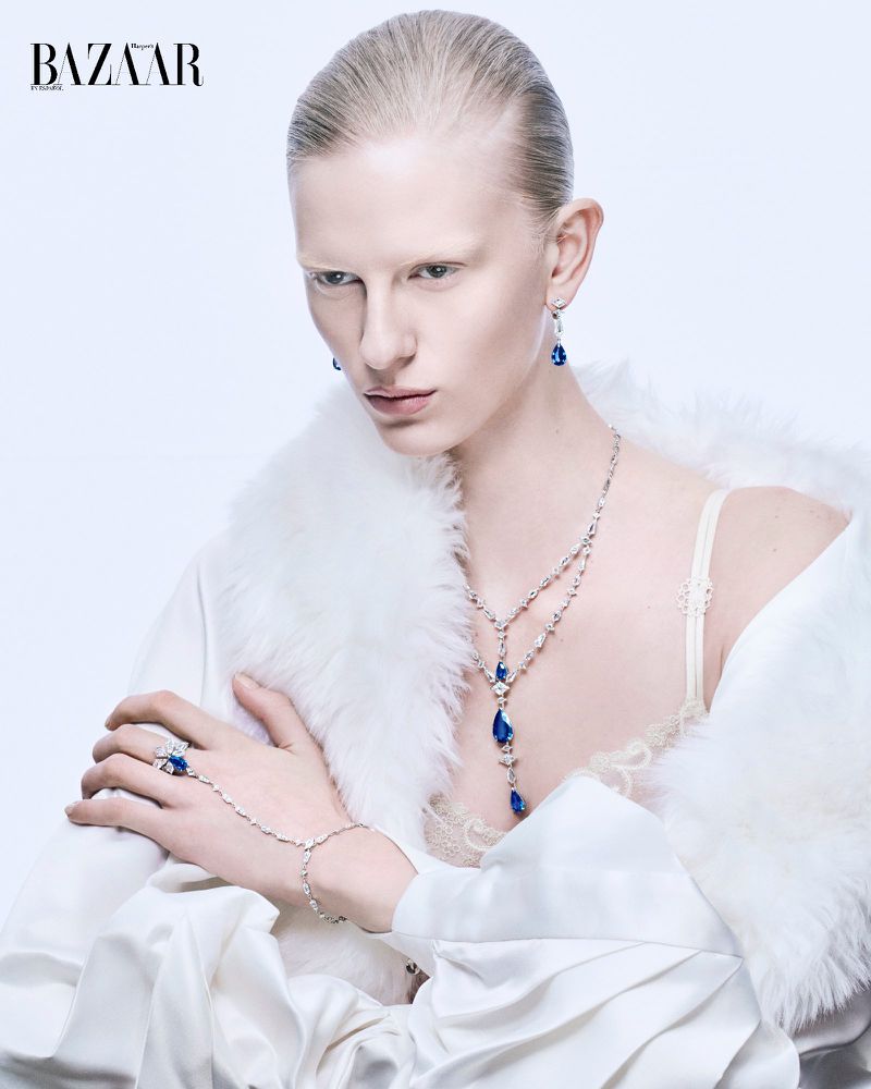Elegant woman with fair skin and white hair poses with hand jewelry, necklace, and earrings against a clean, bright backdrop.