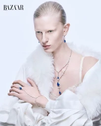 Model wearing fine jewelry and white fur-trimmed coat against a plain background