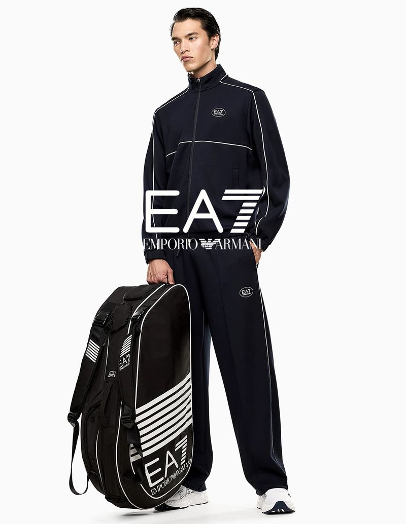 Model wearing a navy EA7 tracksuit and white sneakers while holding a large black sports bag against a plain white background