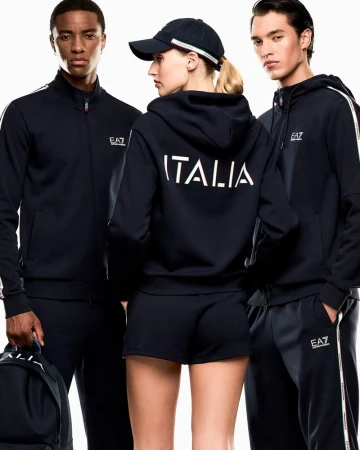 Three athletic models in navy EA7 tracksuits and baseball caps, with 'Italia' text, pose in a bright studio setting for a fashion editorial.