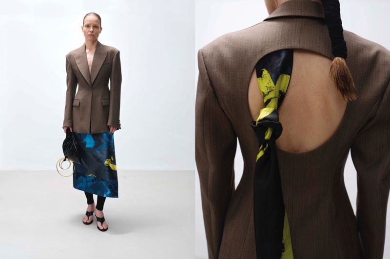Model in a structured brown blazer and patterned blue skirt standing against a plain white studio background.
