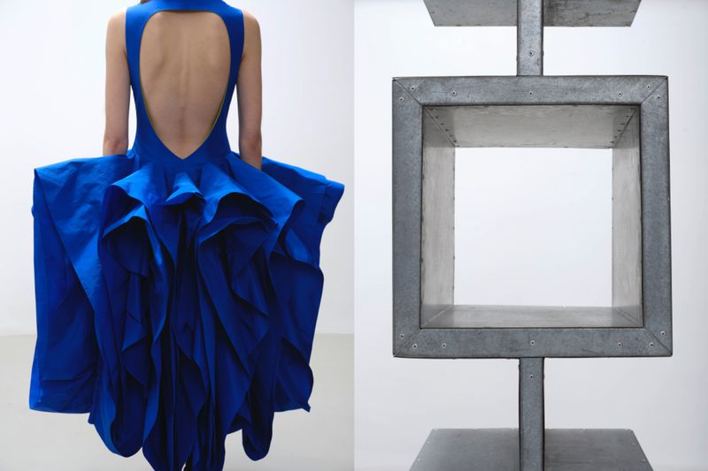 Back view of a model in a vibrant blue Roksanda dress with a cutout back and ruffled skirt beside a metal structure