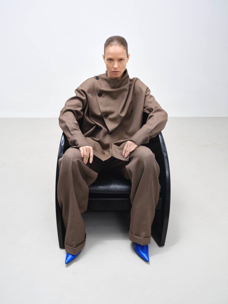 Fashion portrait of an adult woman in a brown suit seated in a black chair against a white background, wearing blue heels.