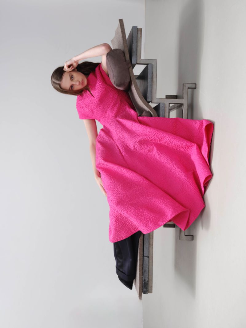Model wearing a vibrant pink textured midi dress posing against a sculptural grey stair structure in a studio setting.
