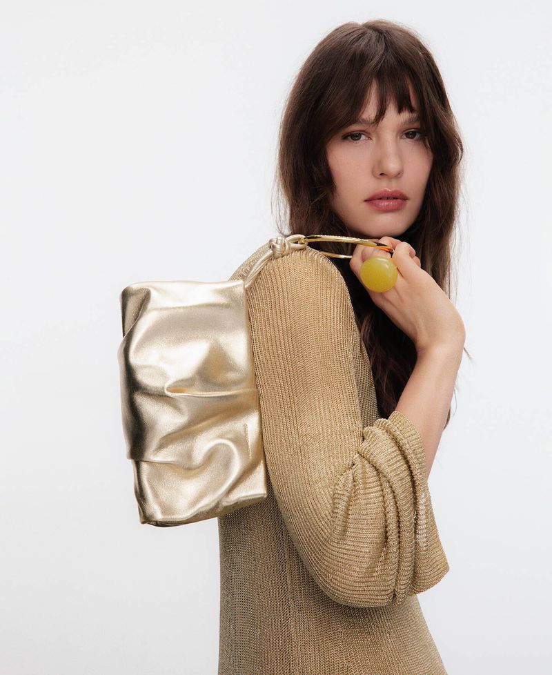 Model wearing a gold textured top holding a metallic gold shoulder bag against a plain white studio background.