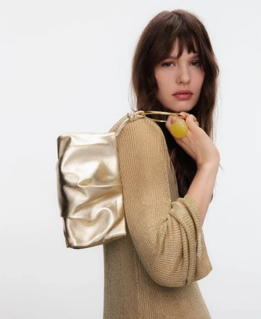 Model wearing a gold textured top holding a metallic gold shoulder bag against a plain white studio background.
