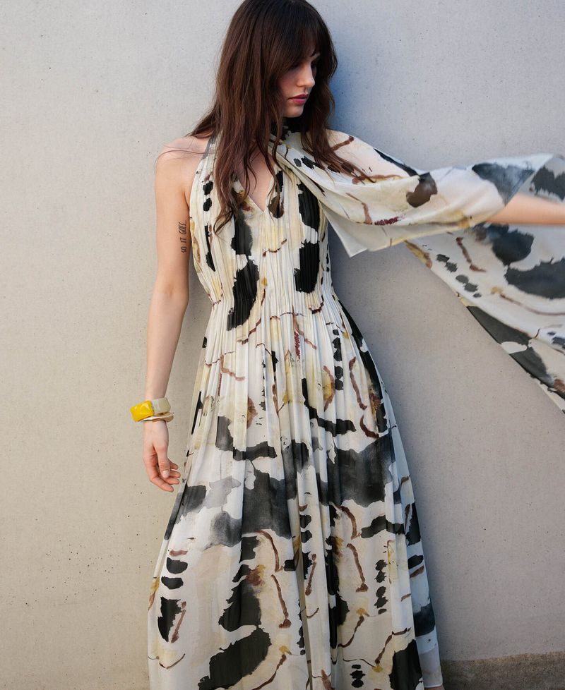 Model wearing a printed halter-neck long dress with a flowing scarf against a plain textured wall